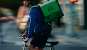 Entregador da Uber Eats em uma bicicleta atravessando a rua em ambiente urbano, com mochila térmica verde nas costas.