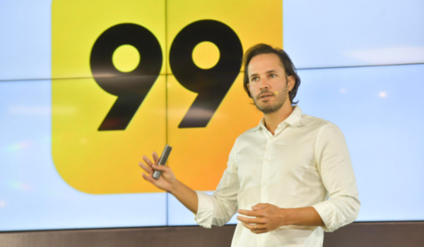 Homem com camisa branca faz apresentação diante de um telão exibindo o logotipo amarelo com o número 99.