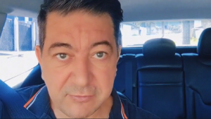 Homem de cabelo escuro e curto, com expressão séria, usando cinto de segurança e camisa azul marinho com detalhe laranja, sentado no banco do motorista dentro de um carro durante o dia.
