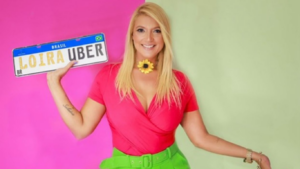Mulher loira sorridente segura uma placa estilizada com os dizeres “Loira Uber” em frente a um fundo dividido entre rosa e verde claro. Ela veste blusa rosa decotada, cinto verde e colar de flor amarela.