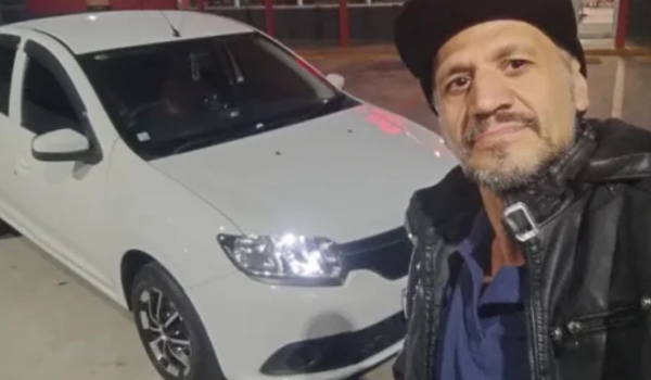 Homem de boné e jaqueta de couro faz selfie ao lado de um carro branco estacionado.