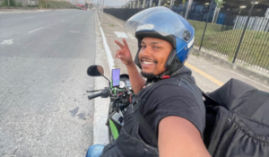 Pessoa sorrindo em uma moto com capacete, fazendo um gesto de paz com a mão, enquanto está em uma estrada.
