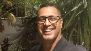 Homem sorridente de óculos e camiseta escura tira selfie à noite em um jardim com folhagens ao fundo.