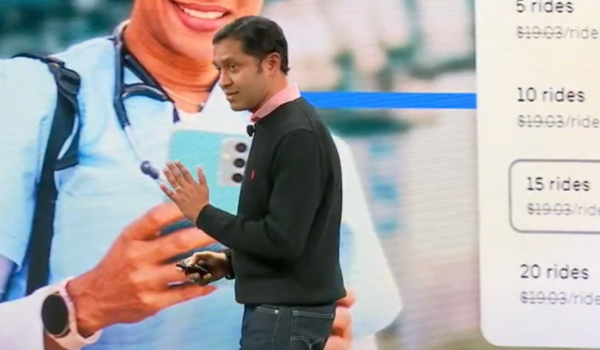 Homem em apresentação ao vivo, gesticulando enquanto fala em frente a uma tela com imagem ampliada de uma profissional da saúde segurando um smartphone.