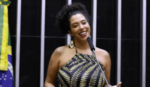 Mulher sorridente discursando em tribuna, com vestido estampado e microfone à frente, diante da bandeira do Brasil.
