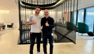 Dois homens sorrindo e posando juntos em um ambiente corporativo moderno, com escada de design contemporâneo ao fundo.