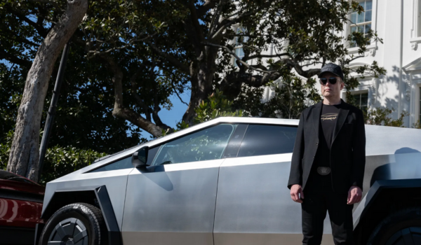 Elon Musk vestindo roupas escuras e boné preto em pé ao lado de um carro Tesla Cybertruck estacionado em uma rua arborizada.