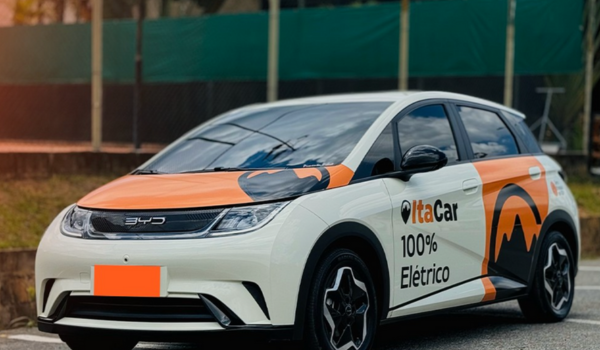 Carro elétrico branco da marca BYD com identidade visual da ItaCar, estacionado em via urbana com cercas e árvores ao fundo.