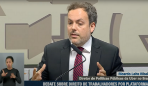 Homem de terno e gravata vermelha fala ao microfone em audiência pública, com intérprete de Libras visível no canto inferior esquerdo.