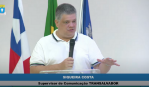Siqueira Costa, supervisor de comunicação da Transalvador, fala ao microfone durante audiência pública. Ao fundo, aparecem as bandeiras da Bahia, do Brasil e de Salvador.