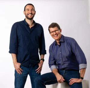 Dois homens sorridentes posam em estúdio com fundo branco; um está de pé com as mãos nos bolsos e o outro sentado em um bloco, ambos vestindo camisa social azul e calça jeans.