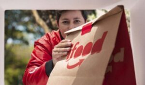 Entregador do iFood visto de dentro de uma caixa ao entregar um saco de papel com o logotipo da marca.