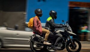Mototaxista conduz passageiro de mochila amarela em uma motocicleta, ambos usando capacetes, em uma rua urbana com fundo borrado por movimento.