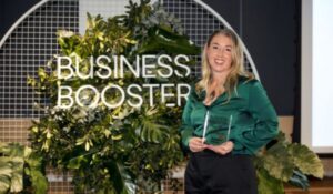 Mulher segurando um troféu transparente em frente a um letreiro com a frase "Business Booster", com fundo decorado por plantas tropicais verdes.