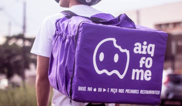 Entregador visto de costas vestindo camiseta branca e capacete, carregando uma mochila de entregas roxa da plataforma "Aiqfome", com logo branco e frase promocional incentivando o download do aplicativo.