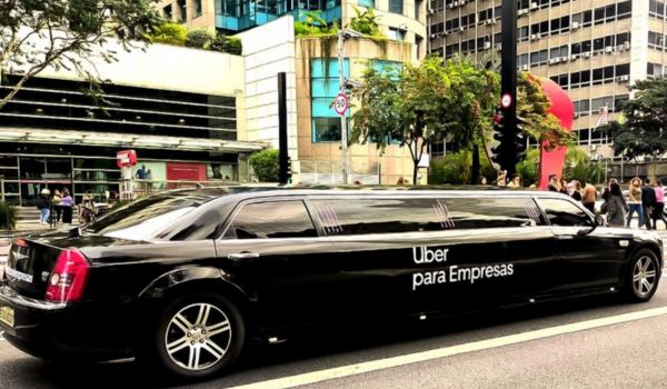 Limusine preta estacionada na Avenida Paulista, em São Paulo, com a frase “Uber para Empresas” escrita na lateral. Ao fundo, pessoas caminham na calçada em frente a edifícios comerciais.