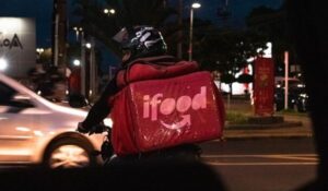 Entregador de moto com mochila vermelha do iFood nas costas trafega à noite por uma avenida movimentada.