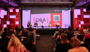 Auditório com várias pessoas assistindo a um painel com cinco mulheres sentadas no palco, participando de uma sessão de perguntas e respostas (Q&A). Um grande telão ao fundo exibe a palavra "Q&A", um QR code e elementos gráficos coloridos.