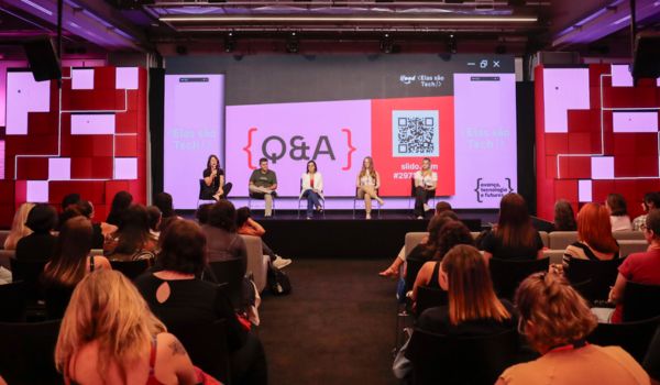Auditório com várias pessoas assistindo a um painel com cinco mulheres sentadas no palco, participando de uma sessão de perguntas e respostas (Q&A). Um grande telão ao fundo exibe a palavra "Q&A", um QR code e elementos gráficos coloridos.