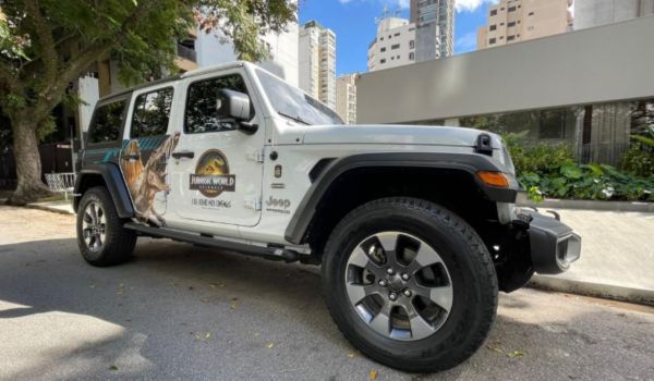 Jeep branco estacionado em rua urbana, com adesivo do filme Jurassic World na porta, mostrando um dinossauro e o logotipo oficial da franquia.