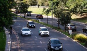 Vista aérea de uma avenida urbana arborizada com três faixas em cada sentido, onde diversos carros circulam em ambos os lados. Há canteiros centrais com grama e árvores, além de calçadas ladeadas por vegetação. Um ciclista aparece na lateral esquerda da via.