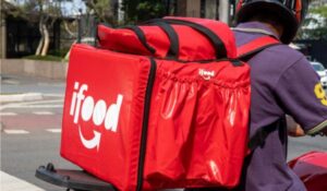 Entregador de moto com mochila térmica vermelha da empresa iFood presa às costas, parado em uma rua da cidade.