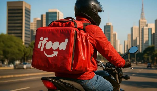 Entregador do iFood de moto, com mochila vermelha, pilotando em uma avenida de cidade grande ao entardecer.