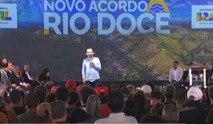 Presidente Lula discursa em evento público diante de uma plateia, com o palco decorado pelo painel "Novo Acordo Rio Doce" ao fundo. Ele usa chapéu branco, camisa azul clara e calça escura, enquanto segura o microfone e fala ao público.