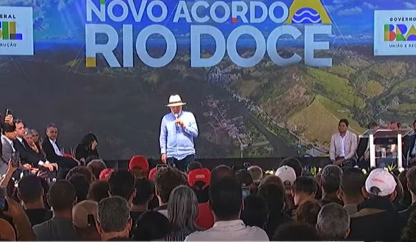 Presidente Lula discursa em evento público diante de uma plateia, com o palco decorado pelo painel "Novo Acordo Rio Doce" ao fundo. Ele usa chapéu branco, camisa azul clara e calça escura, enquanto segura o microfone e fala ao público.