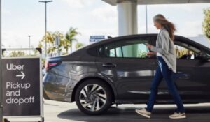 Mulher caminhando em direção à porta traseira de um carro sedan cinza escuro, segurando um celular. Ao lado, uma placa com a marca Uber indica a área de embarque e desembarque com uma seta para a direita.