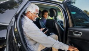 Homem idoso com cabelo grisalho e óculos sorri enquanto entra em um carro preto, segurando um celular. Uma mulher ao volante, também sorridente, o observa do banco do motorista.