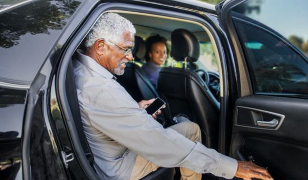 Homem idoso com cabelo grisalho e óculos sorri enquanto entra em um carro preto, segurando um celular. Uma mulher ao volante, também sorridente, o observa do banco do motorista.