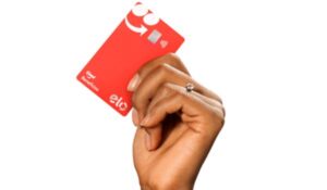 Mão segurando cartão vermelho de benefícios do iFood com bandeira Elo e pagamento por aproximação.