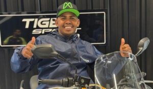 Homem sorridente vestindo jaqueta azul e boné preto com detalhes verdes, posando sobre uma motocicleta, com os dois polegares levantados. Ao fundo, há uma placa com o texto “TIGER SPORT”.