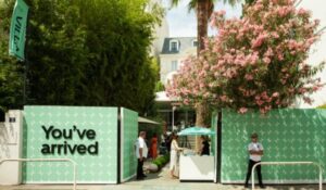 Entrada de um espaço ao ar livre com painéis verdes decorados com padrão geométrico branco e a frase "You've arrived" (Você chegou). Duas pessoas estão paradas próximas ao portão, e há uma árvore florida e uma tenda listrada ao fundo. O ambiente transmite uma atmosfera tropical e acolhedora.