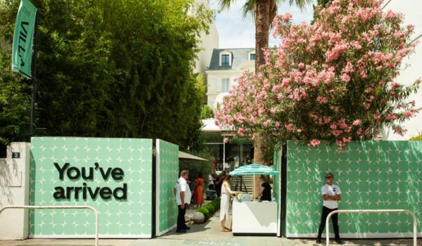 Entrada de um espaço ao ar livre com painéis verdes decorados com padrão geométrico branco e a frase "You've arrived" (Você chegou). Duas pessoas estão paradas próximas ao portão, e há uma árvore florida e uma tenda listrada ao fundo. O ambiente transmite uma atmosfera tropical e acolhedora.