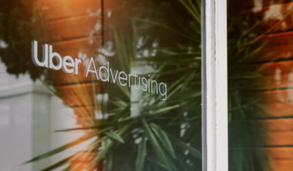 Reflexo de uma planta e parede de madeira em uma vidraça com a inscrição “Uber Advertising” em letras brancas.
