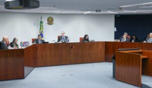 Sessão de julgamento em um tribunal brasileiro, com juízes togados sentados ao redor de uma bancada em formato de "U", bandeira do Brasil e brasão da República ao fundo.