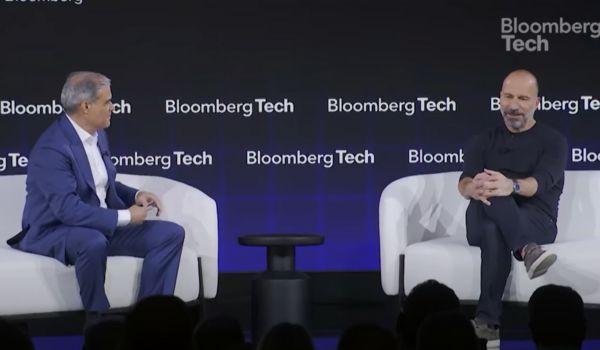 Dois homens sentados em poltronas brancas participam de uma conversa em um palco com fundo preto que exibe repetidamente o logotipo "Bloomberg Tech". O homem à esquerda veste terno azul com camisa branca, enquanto o homem à direita está de preto, com as pernas cruzadas e as mãos unidas.