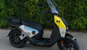 Scooter elétrica preta com detalhes em branco e amarelo, adesivada com o número 99 na parte frontal e os dizeres "100% elétrica" na lateral. A moto está estacionada sobre piso de concreto, com uma parede coberta por plantas verdes ao fundo.