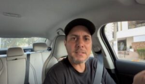 Motorista de aplicativo dentro do carro, usando cinto de segurança, olhando diretamente para a câmera com expressão séria. Ele usa boné preto e camiseta escura. Ao fundo, é possível ver parte do banco traseiro e uma calçada com edifício do lado de fora da janela.