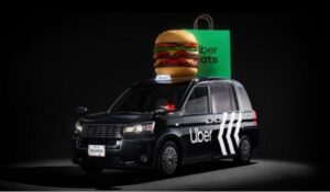 Carro da Uber preto estilizado, com o logotipo da Uber e setas brancas nas portas. Sobre o teto do veículo, há uma escultura gigante de hambúrguer com alface, tomate e queijo, ao lado de uma sacola verde com o logo do Uber Eats.