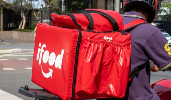 Entregador de moto com mochila térmica vermelha da empresa iFood presa às costas, parado em uma rua da cidade.