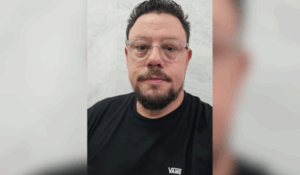 Homem de óculos com armação transparente, barba e camiseta preta da marca Vans, posando de frente para a câmera diante de uma parede de mármore clara.