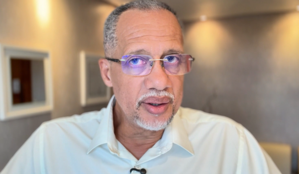Homem de meia-idade com cabelo grisalho e óculos com lentes azuladas, vestindo camisa social clara, fala diretamente para a câmera em ambiente interno bem iluminado.