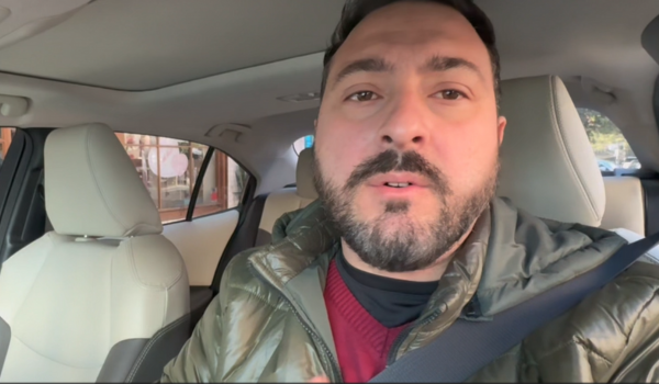 Homem com barba, vestindo jaqueta verde e suéter vermelho, fala diretamente para a câmera enquanto está dentro de um carro.