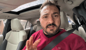 Homem de cinto de segurança afivelado, sentado no banco do motorista de um carro moderno, com teto solar aberto. Ele usa um suéter vermelho e faz um gesto com a mão, como se estivesse explicando algo.