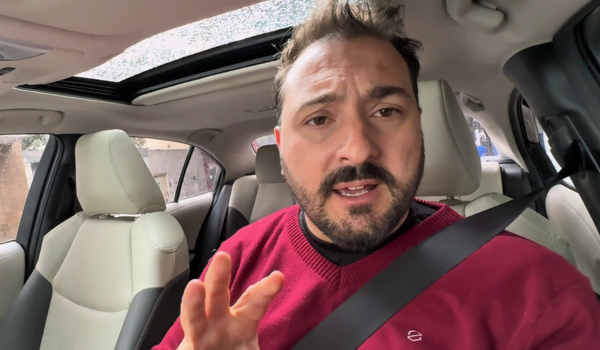 Homem de cinto de segurança afivelado, sentado no banco do motorista de um carro moderno, com teto solar aberto. Ele usa um suéter vermelho e faz um gesto com a mão, como se estivesse explicando algo.