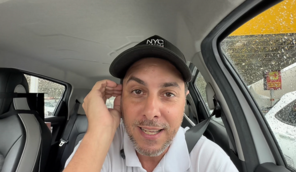 Homem dentro de um carro em dia chuvoso, usando boné preto com a inscrição "NYC Store", camisa polo branca e cinto de segurança, olhando para a câmera com expressão amigável.