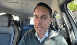 Homem de cabelo curto e barba rala, usando camisa branca e casaco verde-escuro, fala diretamente para a câmera de dentro de um carro.